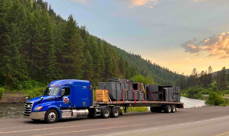 A flatbed truck carries freight through a hilly forest region by a lake