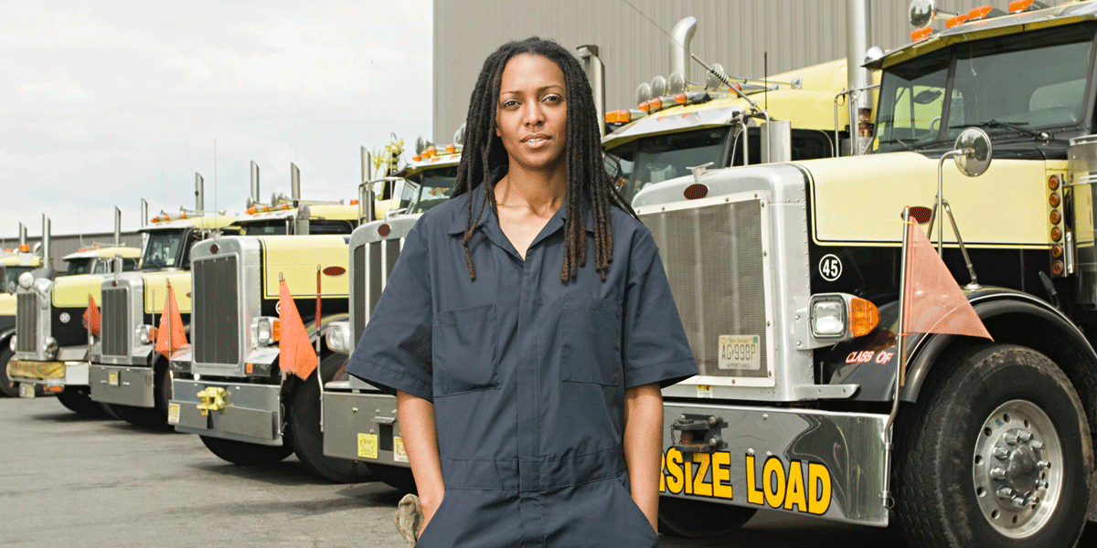 Female diesel mechanic in coveralls standing in front of a line of semi-trucks. 