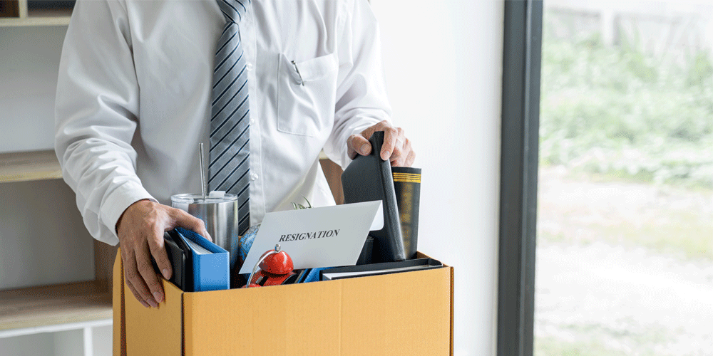 Man in white button down and tie holding a box with his packed-up desk in it. A white piece of paper in the box says "Resignation."