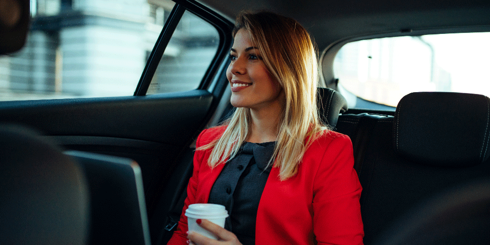 Businesswoman in a red blazer sitting in the back of a taxi with a to-go cup of coffee and a laptop.